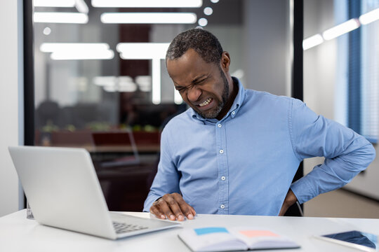 Businessman sitting at desk in modern office experiences back pain while working on laptop. Image highlights discomfort associated with prolonged sitting.