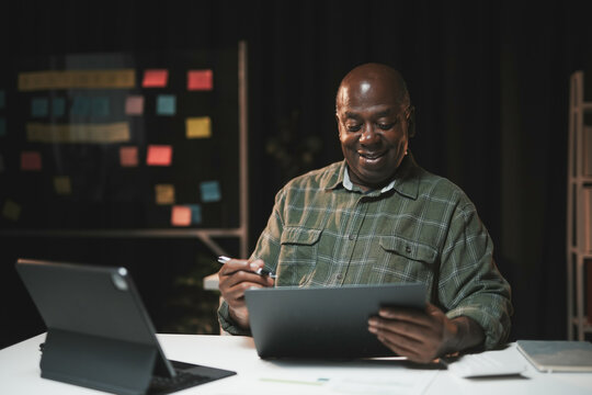Senior black man is working late and using a digital tablet with a stylus, smiling at the screen