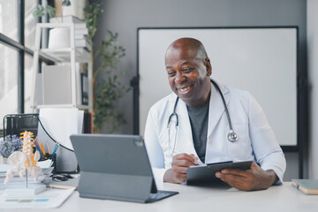 Doctor is smiling while having a video call on his tablet and taking notes on a clipboard