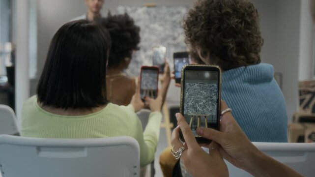Medium full rear shot of multiethnic guests sitting on chairs during lecture in gallery, taking photos of abstract masterpiece on smartphones, while male art curator is presenting it to public