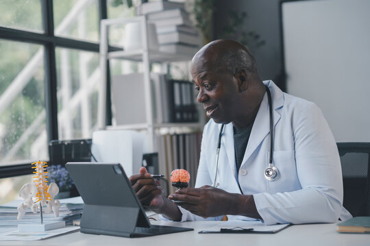 Senior doctor is using a human brain anatomical model while explaining something to a patient during a video call on a tablet