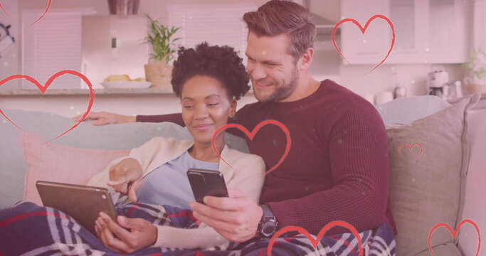 Image of hearts and light spots over diverse couple using smartphone and tablet on sofa