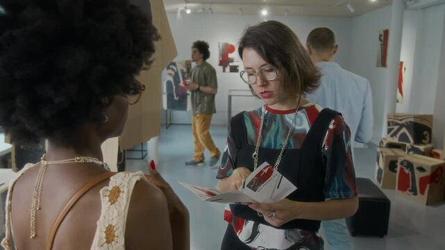 Medium shot of eccentric Caucasian female artist in colourful top, bustier, glasses, holding information booklet, chatting to black female guest about her art vision at personal exhibition in museum