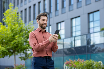 A man in a red shirt is looking at his cell phone. He is smiling. The building behind him is gray