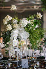 Table number 7. Stands, sign number seven. Setup detail. Luxury reception. Elegant table decorated composition flowers in party area. Beautifully organized event. Wedding hall with atmospheric decor.