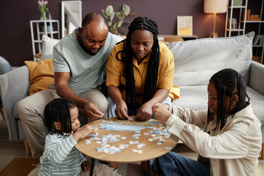 Family gathered around wooden table solving puzzle, focusing on pieces and working together. Living space is warm and inviting, with plants and lamps adding ambiance