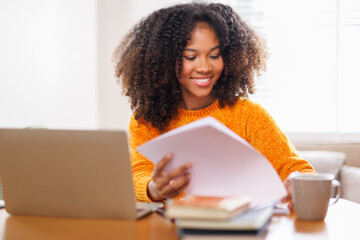 Female digital nomad smiling happily as she types on her laptop at a home. Afro haired woman dressed in casual clothing and working online in a relaxed environment.