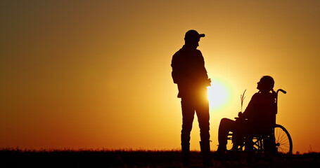 Silhouette of a man assisting a person in a wheelchair against a vibrant sunset backdrop.
