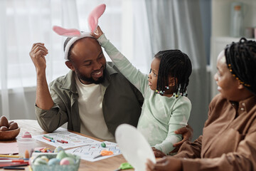 African American man wearing bunny ears sitting at table engaging in Easter craft activity with...