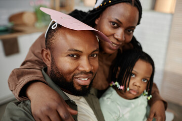 Portrait of smiling family with father wearing paper hat, mother hugging him from behind, and child leaning in closely. All members appearing joyful and relaxed in indoor setting