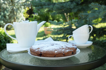 Countryside table setting outdoor in the garden. White porcelain cups, teapot with herbal tea, homemade sweet pie with cherry. Family tradition, beautiful place outside. Side view. 