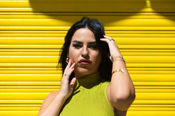 Young Spanish woman, brunette and beautiful with green dress on yellow background touches her hair and face and looks at the camera in a daring and shameless way. Beauty and fashion concept.