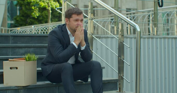 Businessman sitting on stairs near office center with carton box and documents with a table flower as lost job. Male office worker in despair lost job. Unemployment rate growing due pandemic.