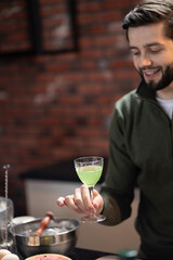 Man with beard smiling while holding green cocktail in glass. Bartender standing in modern kitchen with brick walls, surrounded by various cocktail-making tools and ingredients