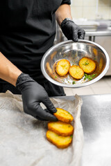 person in black attire and gloves places golden-brown potato slices onto parchment paper. focus is on cooked potatoes, suggesting culinary theme. Ideal for food preparation or cooking-related content.