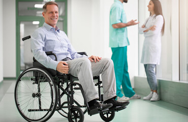 Obraz premium A smiling adult man in a wheelchair sits in a bright hospital hallway. Two medical professionals, converse on the background. The scene suggests recovery, care, and a positive healthcare experience.