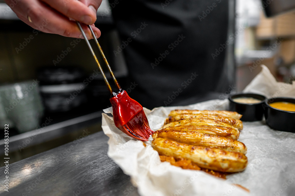 Wall mural Close-up of chef applying red glaze to grilled sausage on parchment paper, with various sauces in the background. The vibrant colors and focus on the culinary action make this image visually appealing