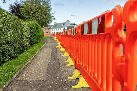 Orange plastic safety barriers separate pavement sidewalk from road during roadworks construction with houses and a hedge in background