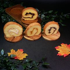 Four soft slices of rolled cake with different fillings, ranging from fruit jam to nut cream, served on wooden boards with a background of green leaves and a yellow-orange autumn leaf.