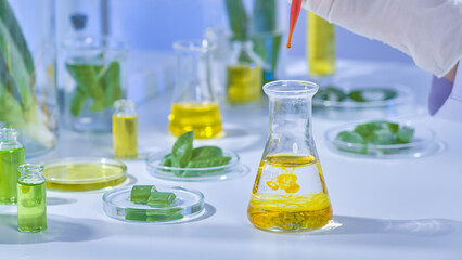 Scientist carefully pours orange liquid into a transparent liquid-filled flask. Aloe samples and other liquids are arranged around the laboratory table. A instance of an aloe vera product photo.
