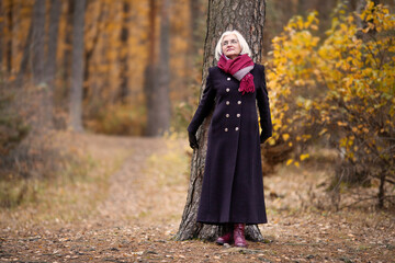 Mature woman standing with tree trunk in autumn forest, dressed in cozy fall attire, black coat and scarf, gazing thoughtfully at vibrant fall foliage. Reflection and connection with nature, wellness