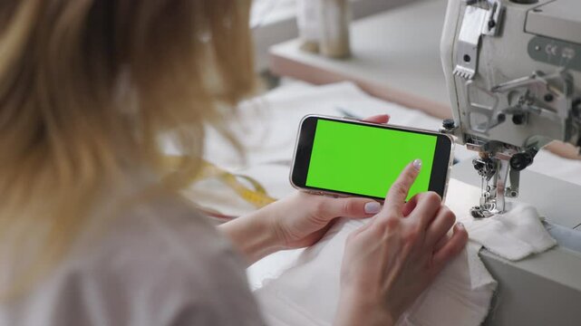 Fashion designer interacts with smartphone that displays green screen, perfect for customizing content. The designer is seated next to sewing machine, implying that she is multitasking.