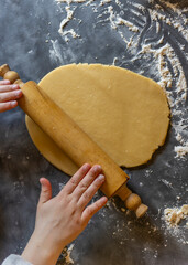 Teenage girl rolls out an oval disc of golden-brown cookie dough with a wooden rolling pin on a dark counter work top covered in flour