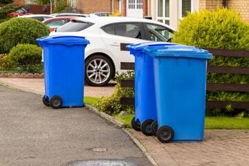 Blue waste recycling wheelie bins trash cans lined up in a row for collection on a residential street with parked cars in driveways