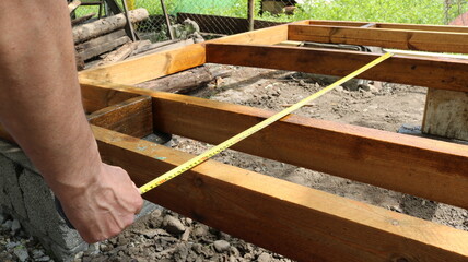 A man holds a tape measure and measures the distance between the wooden beams that are part of the structure and are mounted on concrete piers. 