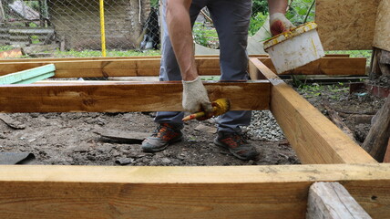 A person wearing work clothes and gloves applies a protective coating or paint to a wooden structure. He holds a brush in one hand and a bucket of varnish or paint in the other.
