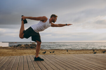 Young sporty athletic toned fit sportsman man wear sports clothes earphones hold leg lean forward spread hand warm up train at sunrise sun over sea beach outdoor on pier seaside in summer day morning.