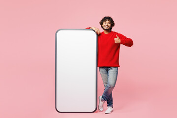 Full body young Indian man he wears red sweater casual clothes big huge blank screen mobile cell phone smartphone with area show thumb up isolated on plain pink background studio. Lifestyle concept.