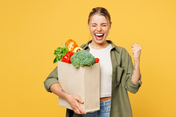 Young woman wear casual clothes hold bag for takeaway mock up with food products do winner gesture celebrate clench fists isolated on plain yellow background. Delivery service from shop or restaurant.