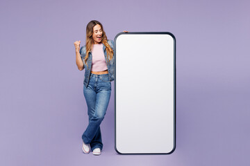 Full body young woman she wear denim jacket violet top casual clothes big huge blank screen mobile cell phone smartphone with area do winner gesture isolated on plain pastel light purple background.