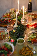 Christmas feast table with plates of food, glasses of wine and champagne, with festive decorations of pinecones, golden stars paper garland and long candles in evergreen foliage centerpieces