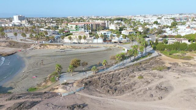 Aerial drone footage showcases the serene Cala Mosca at Playa Flamenca, presenting the harmonious blend of rugged coastal terrain and vibrant urban life along the Orihuela Coast, Spain