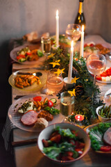 Christmas feast table with plates of food, glasses of wine and champagne, with festive decorations of pinecones, golden stars paper garland and long candles in evergreen foliage centerpieces