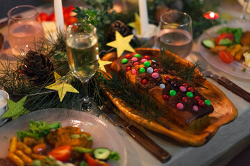 Christmas feast table with plates of food, glasses of wine and champagne, with festive decorations of pinecones, golden stars paper garland and long candles in evergreen foliage centerpieces