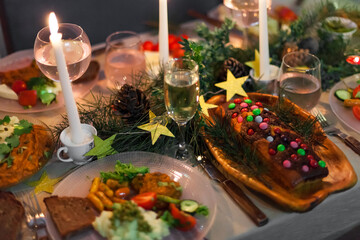 Christmas feast table with plates of food, glasses of wine and champagne, with festive decorations of pinecones, golden stars paper garland and long candles in evergreen foliage centerpieces