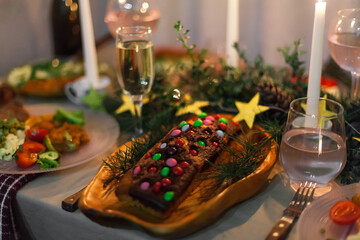 Christmas feast table with plates of food, glasses of wine and champagne, with festive decorations of pinecones, golden stars paper garland and long candles in evergreen foliage centerpieces