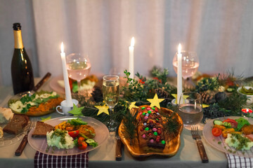 Christmas feast table with plates of food, glasses of wine and champagne, with festive decorations of pinecones, golden stars paper garland and long candles in evergreen foliage centerpieces