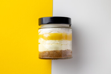 A delicious lemon cheesecake in a glass jar sits elegantly against a vibrant yellow and white split background, highlighting the dessert's beautiful layered presentation.