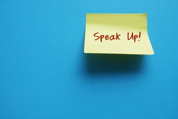 Yellow note stick on copy space blue background with text written SPEAK UP !, to raise voice louder - dare to express opinion frankly and openly , stand up for yourself