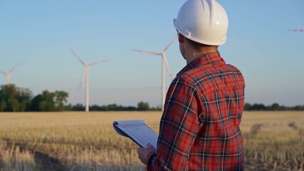 Man engineer, wearing a white protective helmet is taking notes with a clipboard in a field with wind turbines, as the sun sets. Clean energy and engineering concept