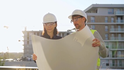 Man and woman architects wearing safety helmets and vests holding blueprint and discussing something on construction site at sunrise