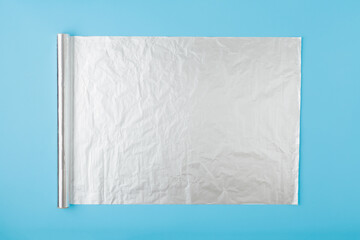 Unroll roll of silver kitchen foil film on light blue table background. Pastel color. Closeup. Top down view.