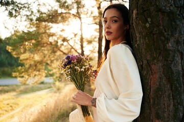 Naklejka premium Near the tree, holding bouquet with flowers. Woman is outdoors in nature with bouquet of flowers