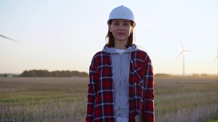 Female engineer is standing straight on a field with wind turbines, as the sun sets. Concept of clean energy and engineering audit