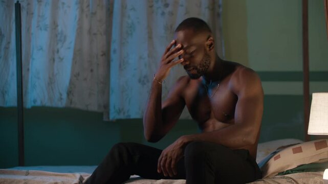 Disappointed and angry shirtless young African man receives bad news on phone whiles sitting on his bed at night. Hits his hands on his bed.