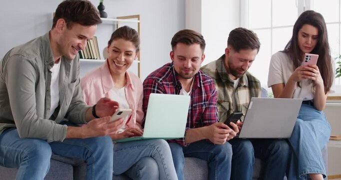 Group of friends using digital devices. Young people sitting on sofa together, working on laptops, using phones, browsing social media, sharing funny memes, laughing, having fun. Technology concept